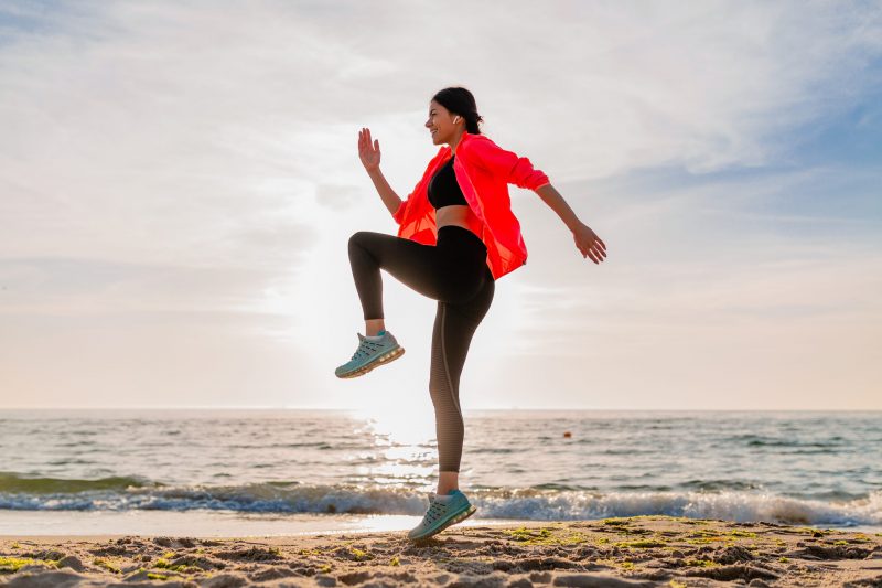 young smiling attractive slim woman doing sports in morning sunrise jumping on sea beach in sports wear, healthy lifestyle, listening to music on earphones, wearing pink windbreaker jacket, having fun young smiling attractive slim woman doing sports in morning sunrise jumping on sea beach in sports wear, healthy lifestyle, listening to music on earphones, wearing pink windbreaker jacket, having fun