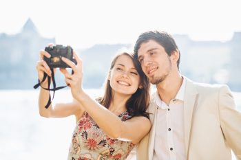 Happy couple in love pose for selfie in modern professional camera, have walk outdoor, explore unknown places in big tourist`s city, model against wonderful landscape with water in background
