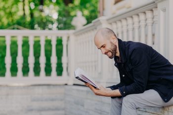 People, hobby and lifestyle concept. Postive bald man reads book with enthusiasm, poses outdoor against white balcony, has happy expression, enjoys calm atmosphere. Middle aged handsome male studies