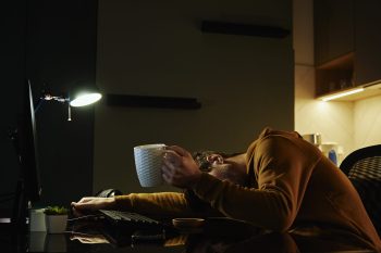 Man sleeping at home office workplace with cup of coffeee in his hand. Freelancer remotely working late. Emotional stress and burnout